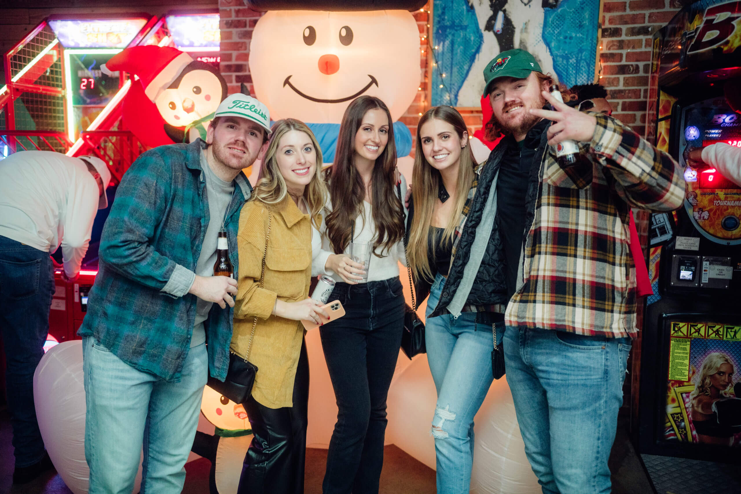 Group of five young adults posing in front of a large inflatable snowman at an arcade with holiday decorations.