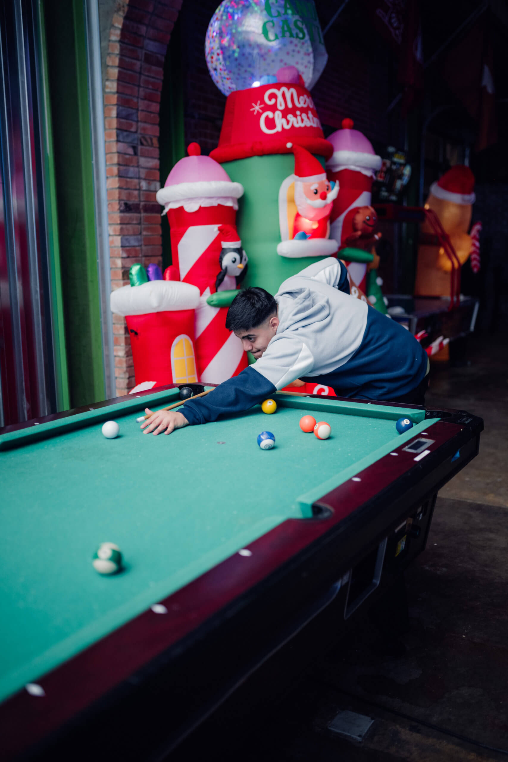 Young man playing pool in a festive arcade decorated with holiday inflatables and Christmas-themed decor.