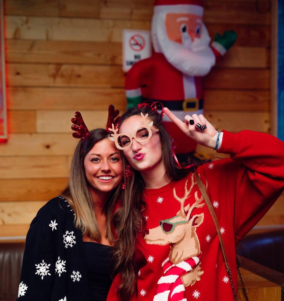 Two women in festive holiday attire pose for a photo with reindeer antlers and Christmas sweaters in front of an inflatable Santa decoration.