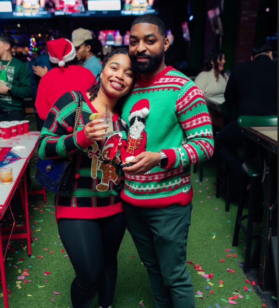 Smiling couple in festive Christmas sweaters pose together at a holiday party, surrounded by colorful confetti and party games.