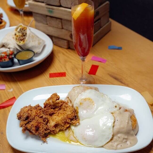 Plate of crispy fried chicken, sunny-side-up eggs, and biscuits smothered in gravy served on a wooden table with a cocktail and Jenga tower.