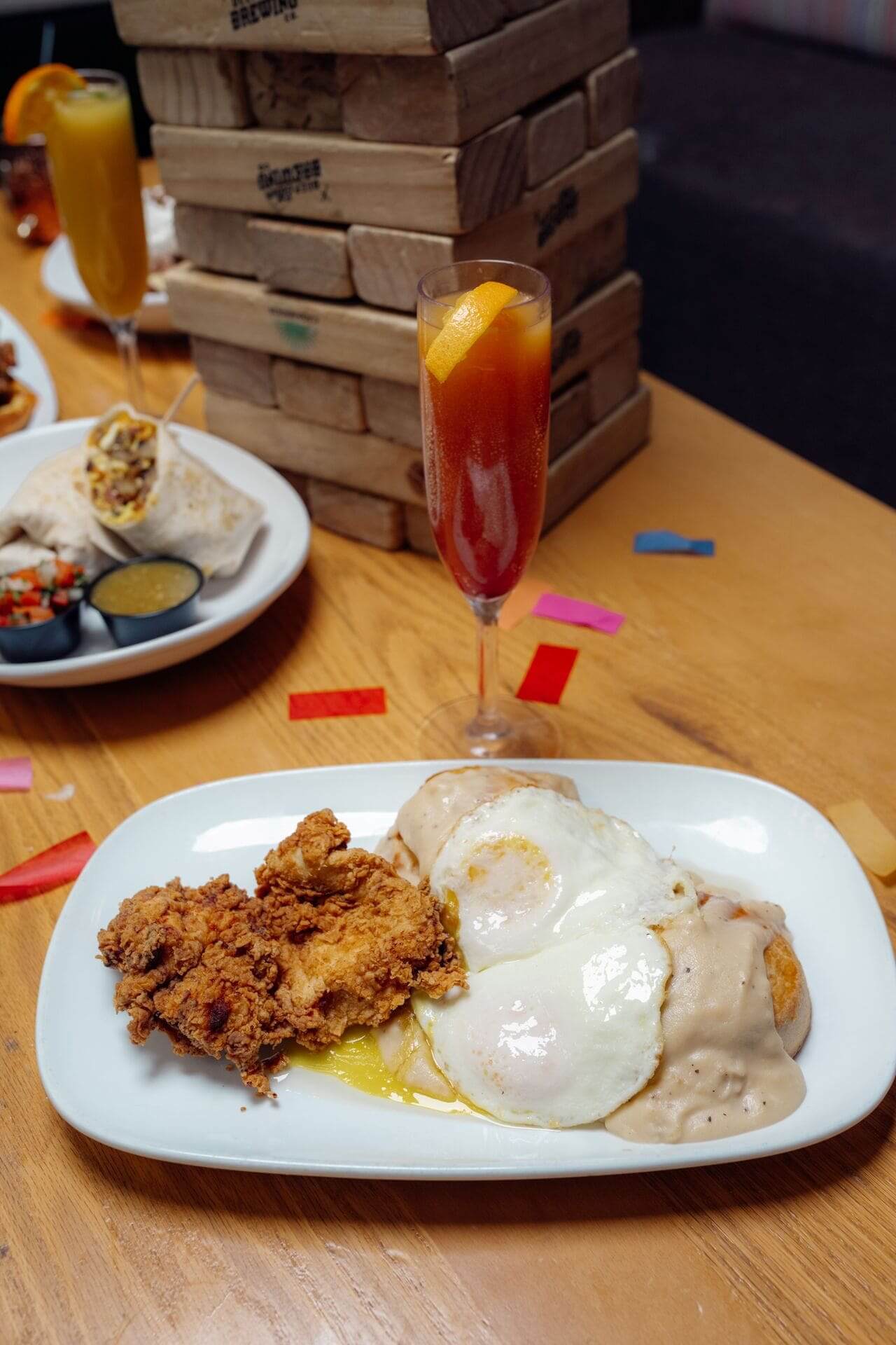 Plate of crispy fried chicken, sunny-side-up eggs, and biscuits smothered in gravy served on a wooden table with a cocktail and Jenga tower.