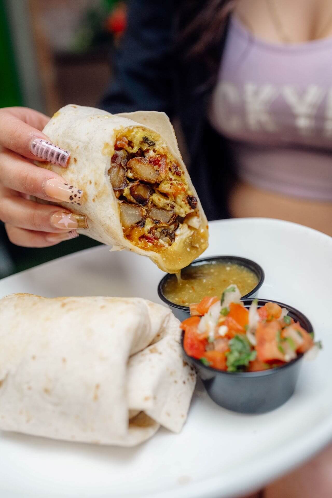 Woman holding a breakfast burrito filled with eggs, potatoes, and vegetables, dipping it into green salsa, with pico de gallo on the side.