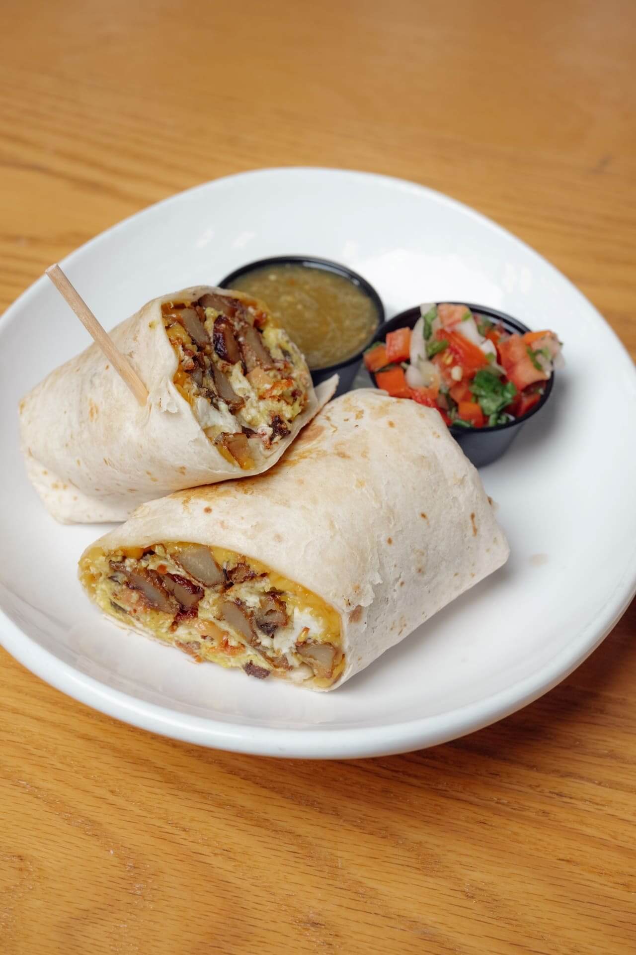 Breakfast burrito cut in half showing eggs, cheese, and potatoes, served with green salsa and pico de gallo on a white plate.