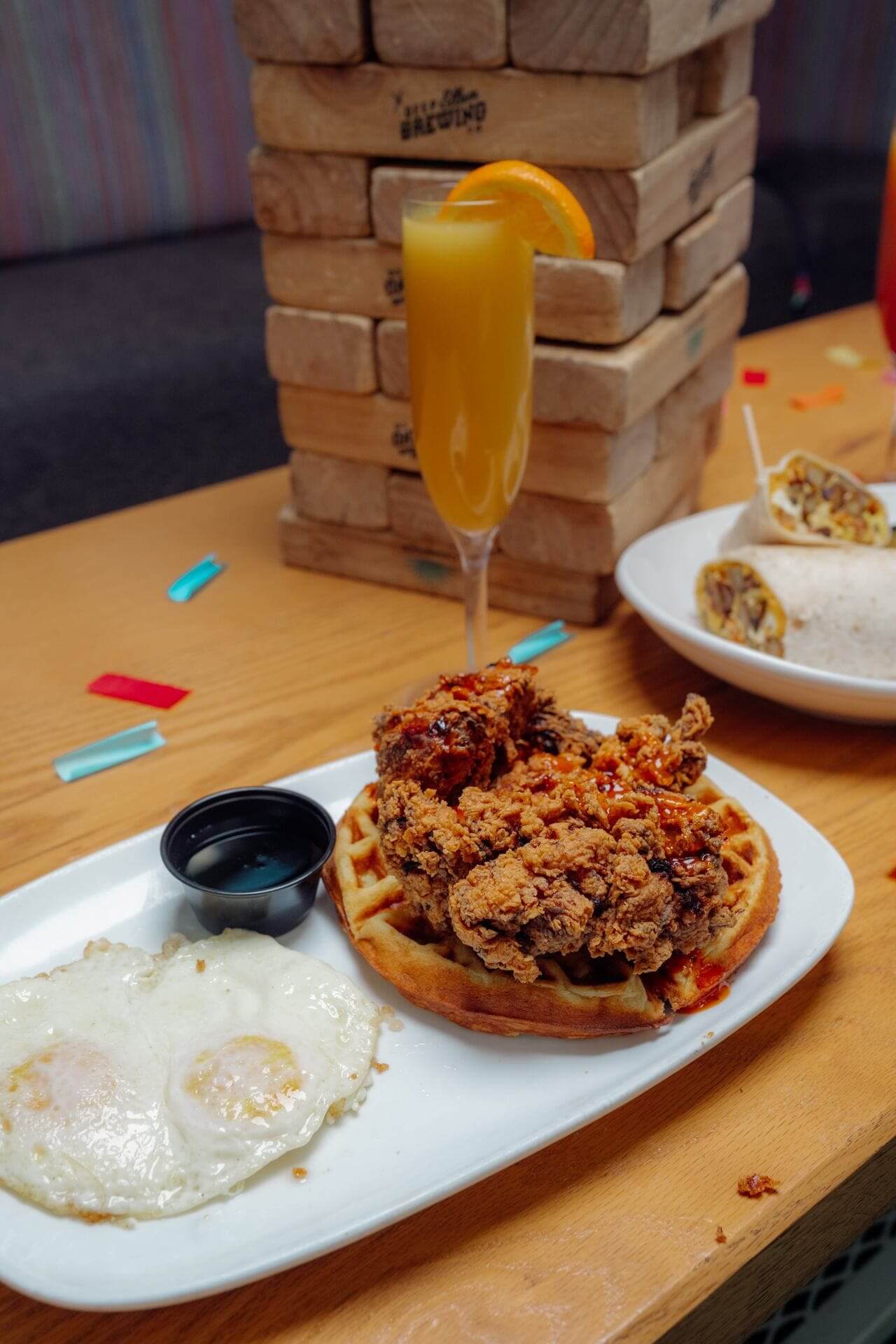 Crispy fried chicken over Belgian waffles served with syrup and fried eggs, with a mimosa and stacked Jenga blocks in the background.
