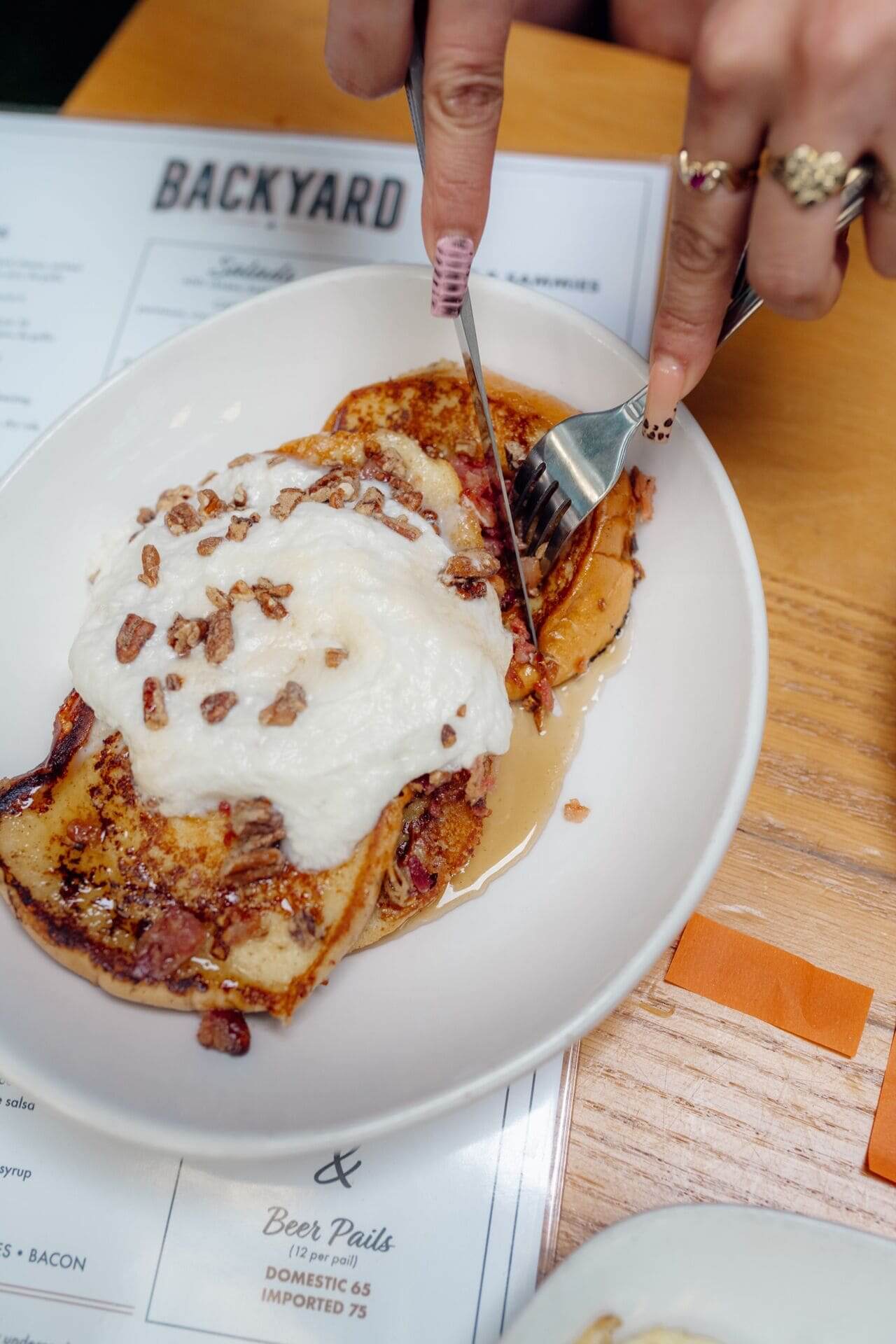 lose-up of French toast topped with whipped cream and chopped pecans being cut with fork and knife at a brunch table