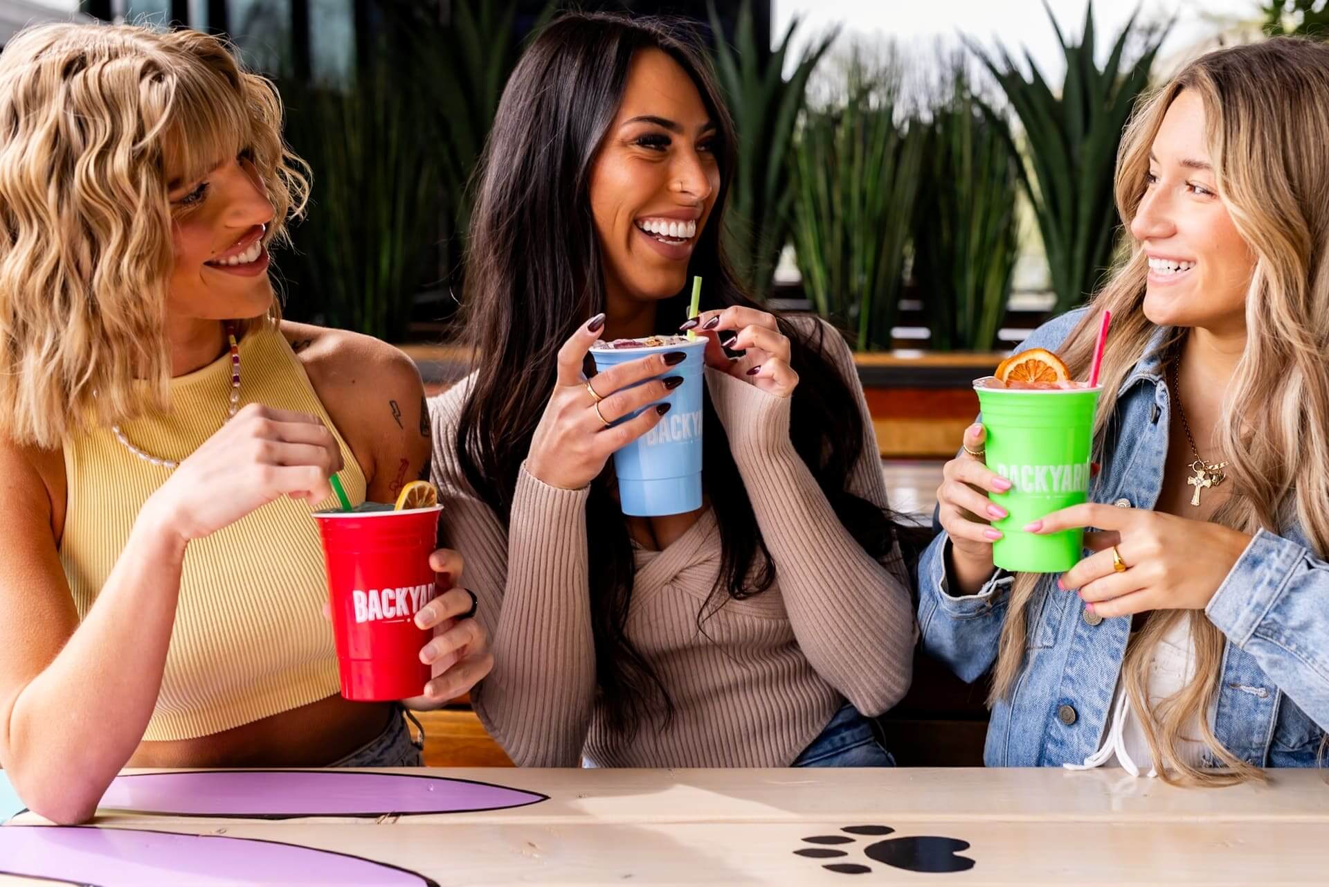 Three women smiling and enjoying colorful cocktails in branded Backyard cups while sitting at a wooden table.