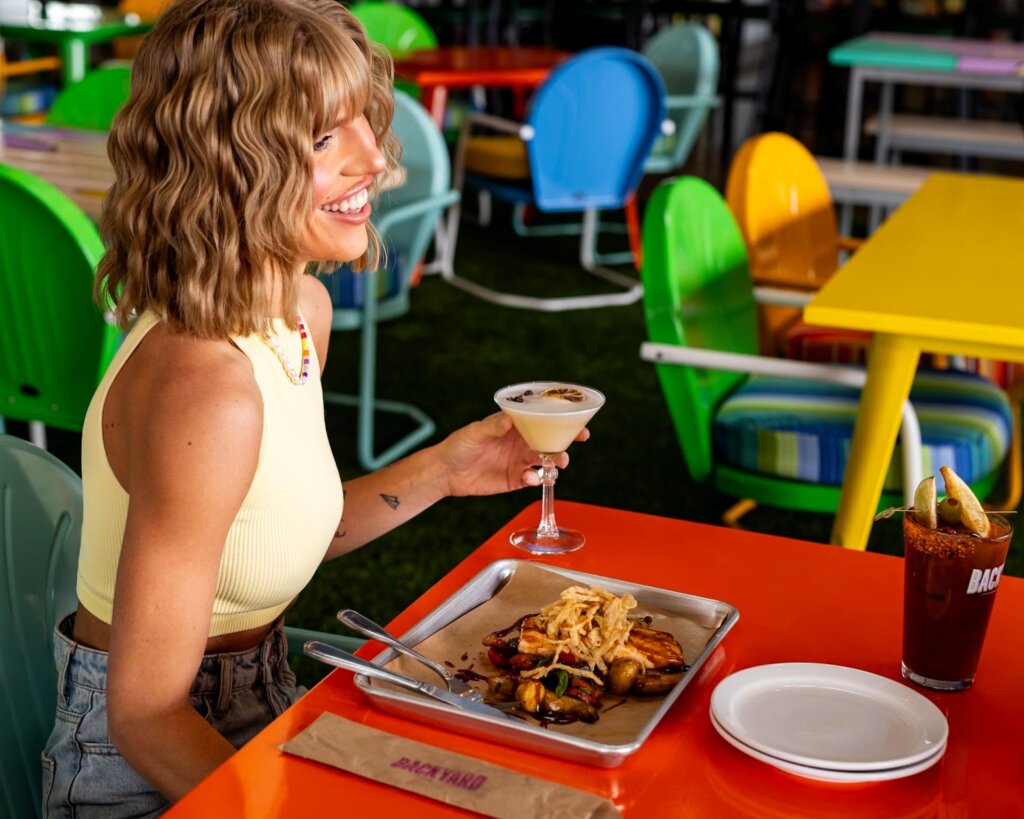Blonde woman holding a cocktail and smiling while sitting at a colorful table with a plate of food and another drink nearby.