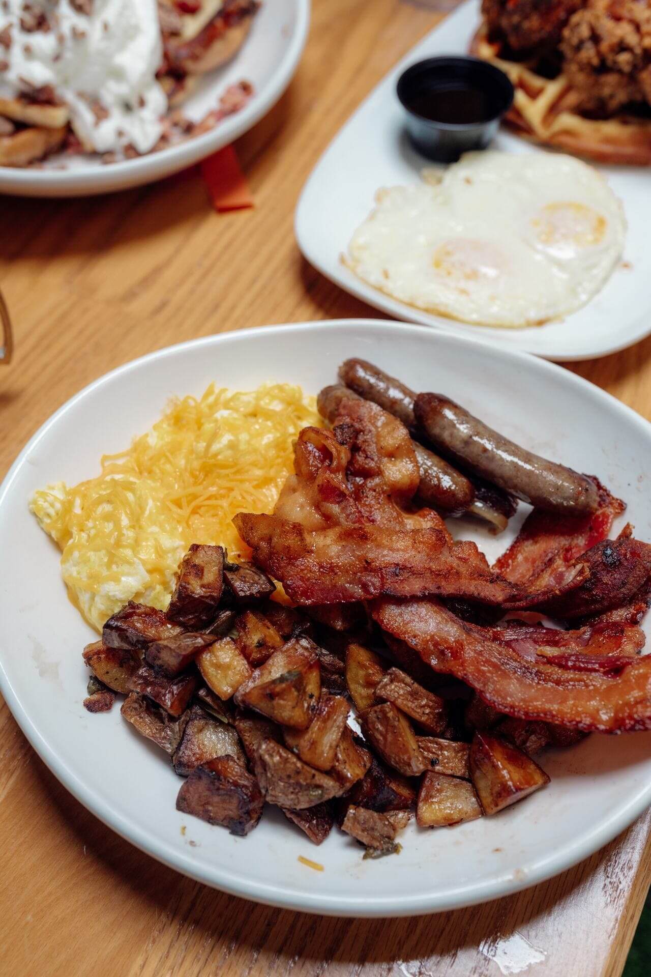 Plate of scrambled eggs with shredded cheese, crispy bacon, breakfast sausage links, and seasoned roasted potatoes.