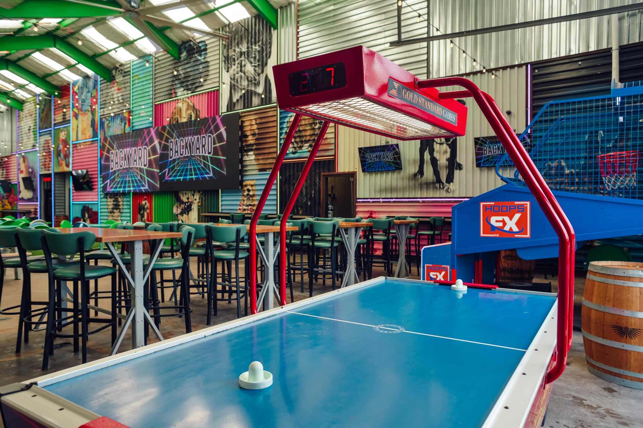 Air hockey table with red scoreboard in a vibrant game-themed bar with neon BACKYARD signage and basketball hoops in the background