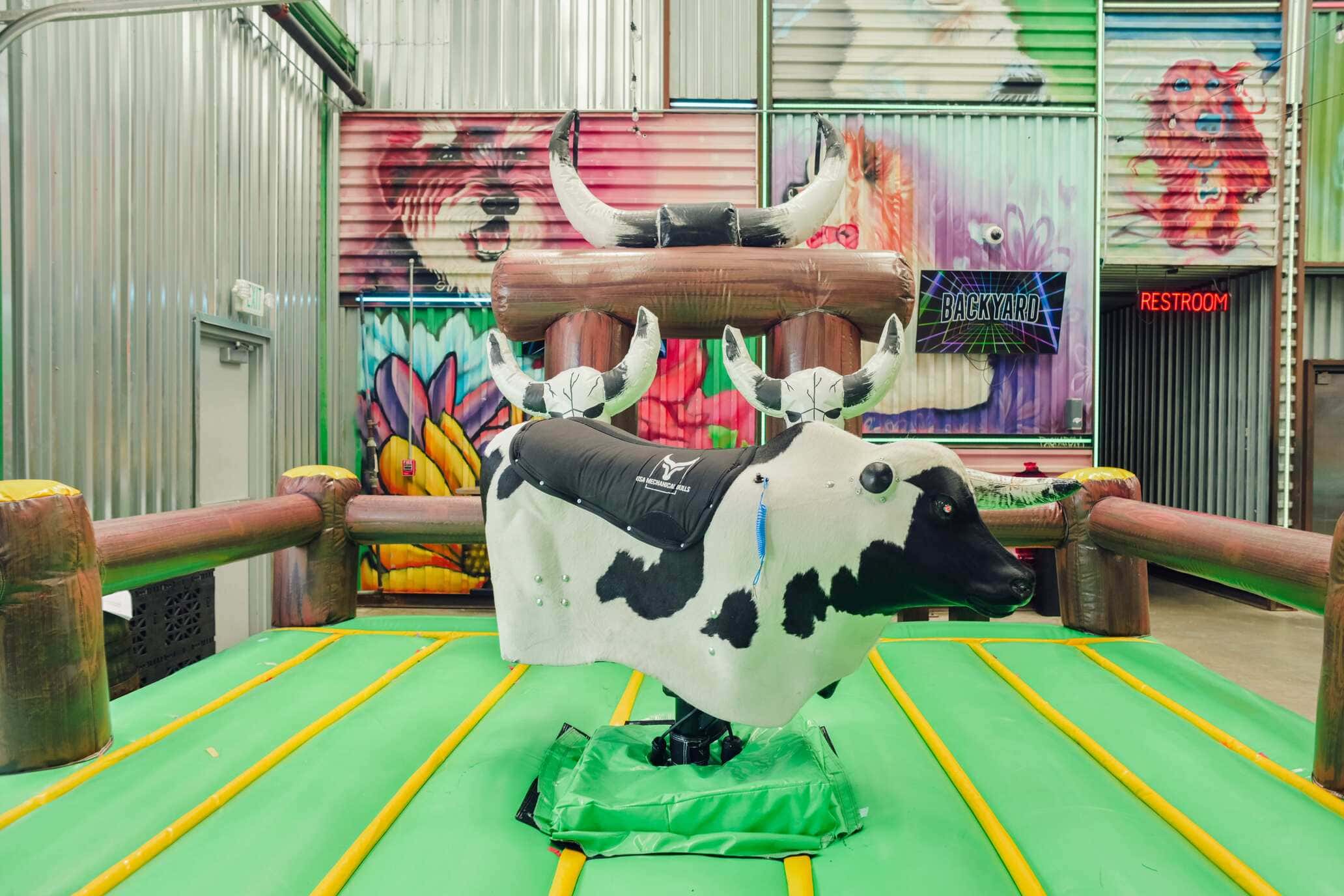 Colorful indoor setup for mechanical bull riding at Backyard Dallas, with a padded green inflatable floor and vivid graffiti-style animal art on metal walls.