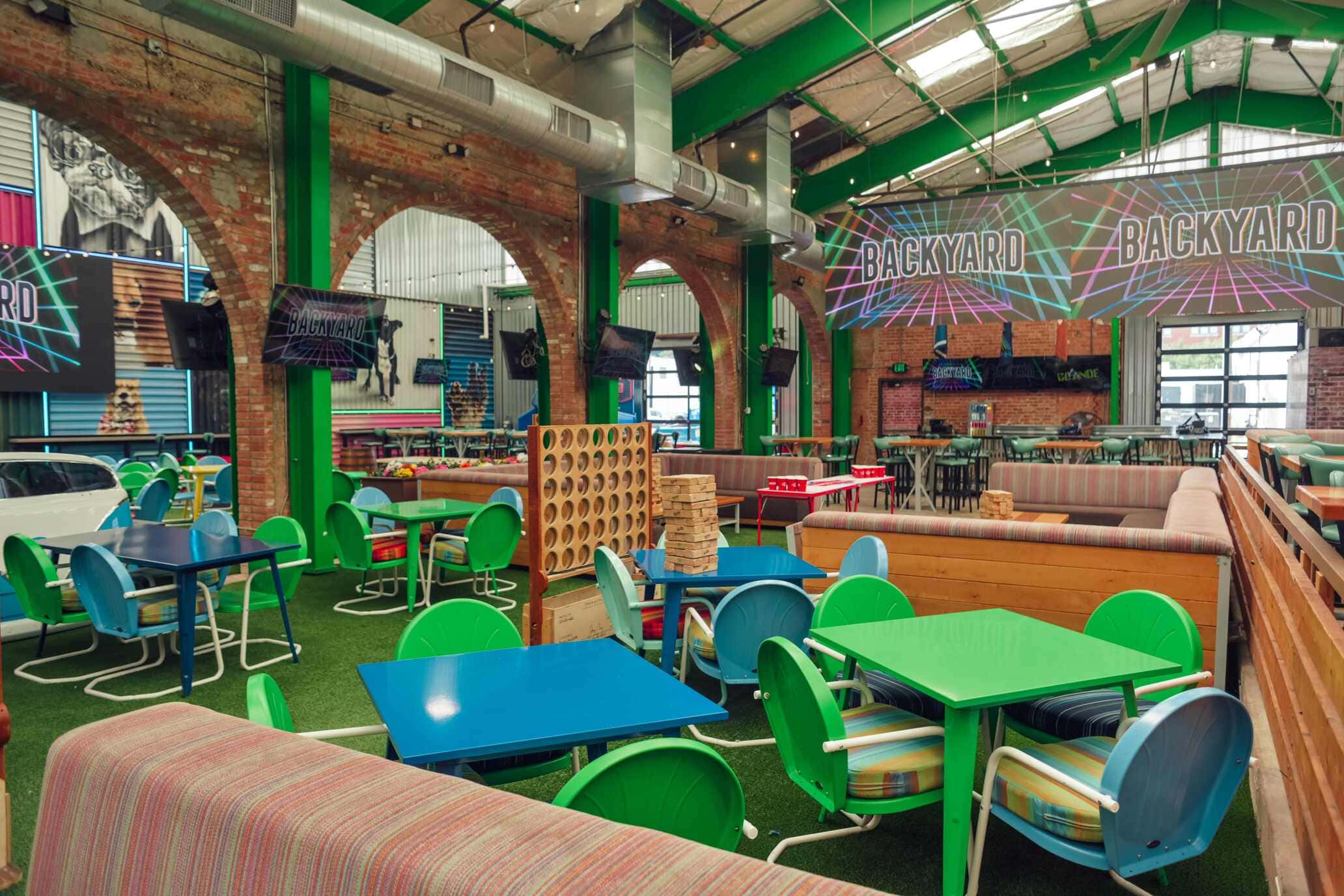 Indoor game lounge featuring colorful tables and chairs, oversized Connect Four and Jenga games, and vibrant "Backyard" signage on TVs beneath a high ceiling with green beams.