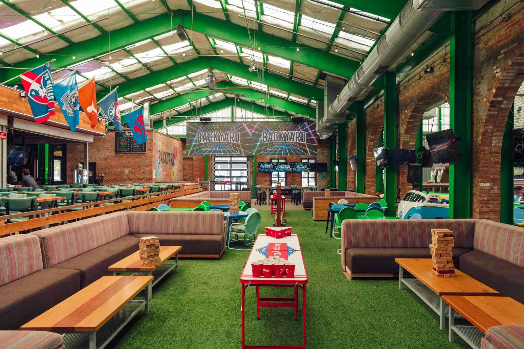 Indoor entertainment venue featuring beer pong, oversized Jenga, and colorful seating under a green metal roof with flags and neon “Backyard” signs.