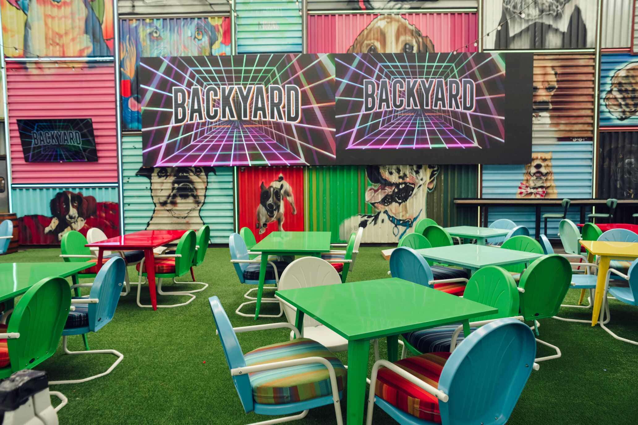Colorful picnic-style dining area with green and blue metal chairs, vibrant green tables, and murals of dogs painted on corrugated metal walls beneath large “Backyard” signs