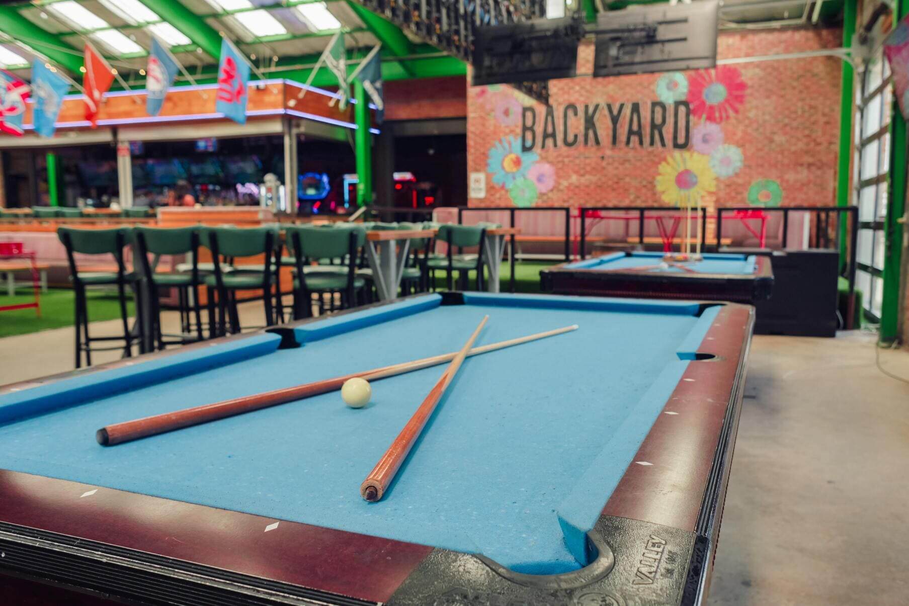 Close-up of pool table with cues and ball, with BACKYARD mural and casual seating in the background.