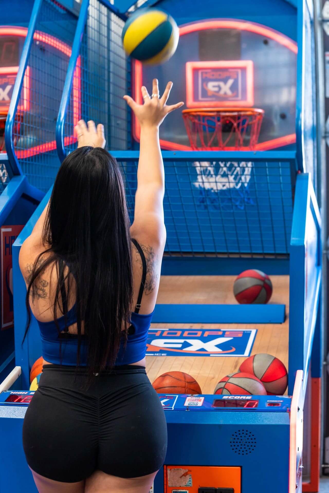 Woman playing arcade basketball, mid-shot, with vibrant neon signage in the background.