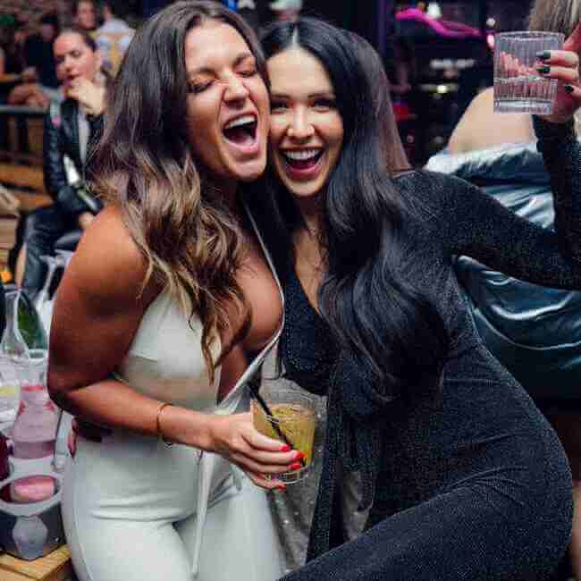 Two women smiling and holding drinks at a lively nightclub celebration.