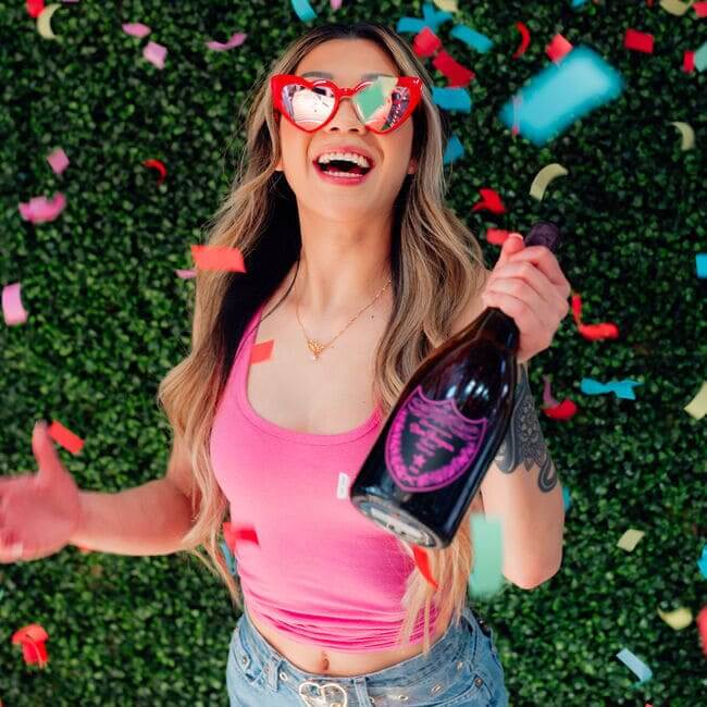 Woman wearing heart-shaped sunglasses celebrating with a champagne bottle as colorful confetti falls.
