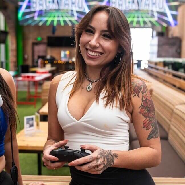 Smiling woman holding a game controller inside a lively bar with arcade games and colorful neon lights in the background.