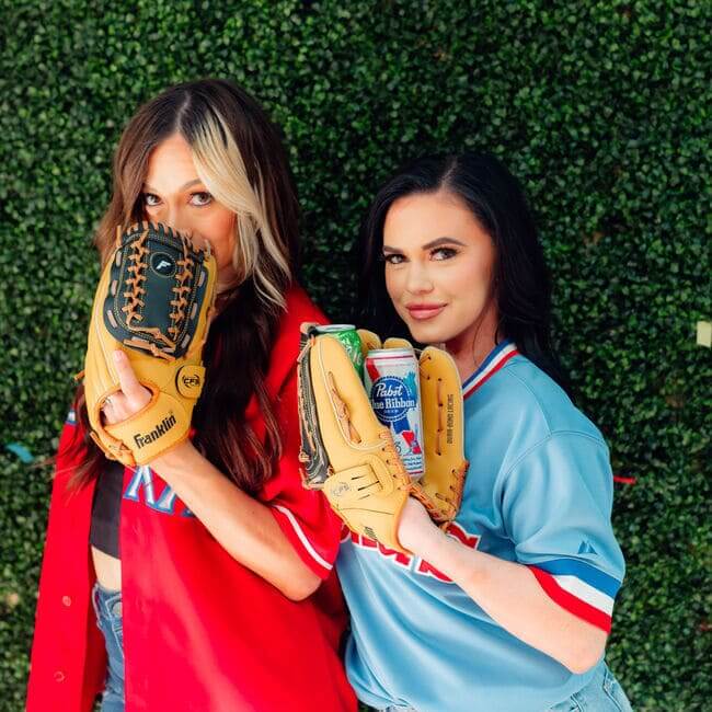 Two women in baseball jerseys holding gloves and a beer can in front of a green hedge
